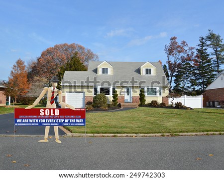 Mannequin wearing red tie holding Real estate sold (another success let us help you buy sell your next home) sign suburban cape cod home autumn blue sky day residential neighborhood USA