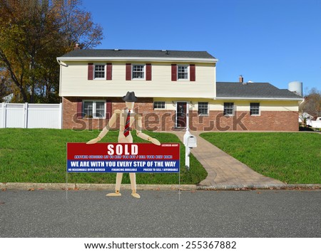 Mannequin wearing red striped tie and black hat holding real estate sold (another success let us help you buy sell your next home) sign suburban home sunny clear blue sky residential neighborhood USA