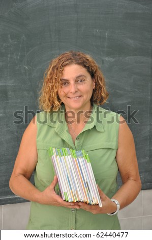 Beautiful Professional Mature Latin School Teacher holding Books in front of Classroom Blackboard