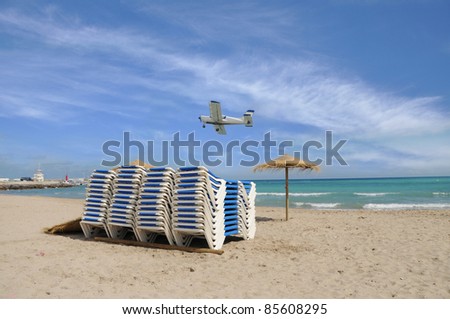 Lounge Chairs Stacked on Sandy Tropical Beach with Umbrella and Small Airborne Plane Flying over Sea Sunny Blue Sky Day