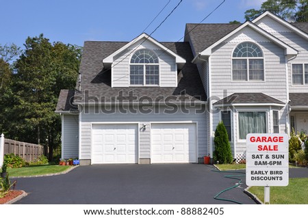 Garage Sale Sign on Black Top Driveway Edge in Front of Suburban Two Car Garage Home Sunny Blue Sky Day