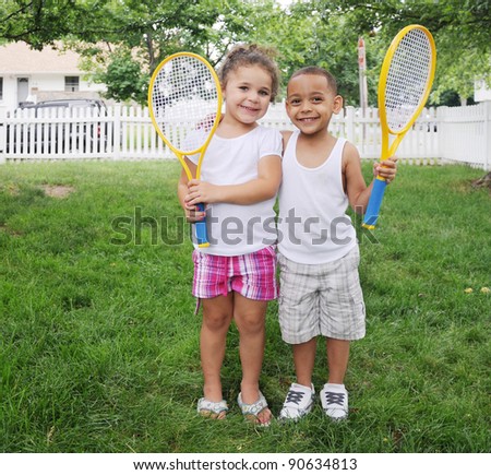 Two Children Holding Tennis Racket Smiling Looking at Camera on Lush Green Suburban Front Yard Lawn Grass