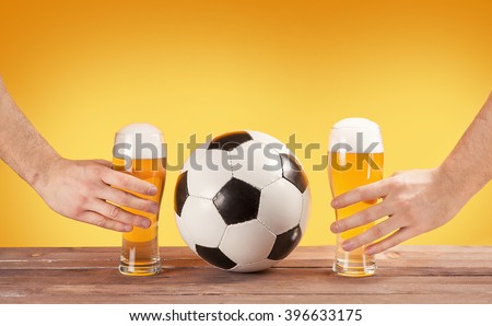 two male hands holding glasses of beer near soccer ball. Yellow background
