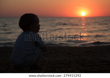 little boy sitting on his haunches and looks like the sun sets on the sea