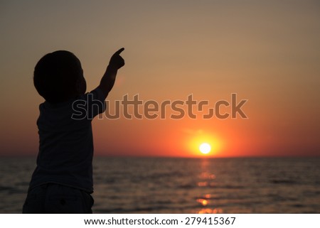 little boy standing back and watching the sun go down over the sea and is pointing at the sun