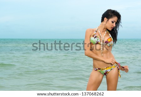Beautiful sexy young woman standing tying her bikini at the seaside with an ocean backdrop and copyspace