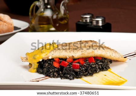 Plate of halibut fillet over a bed of rice with squid ink, capers tomato and mustard in a restaurant setting with copyspace.