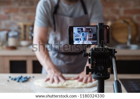 baker online courses. food preparing and culinary training class concept. smiling bearded chef kneading dough in the kitchen and shooting video of himself using mobile phone on a tripod.