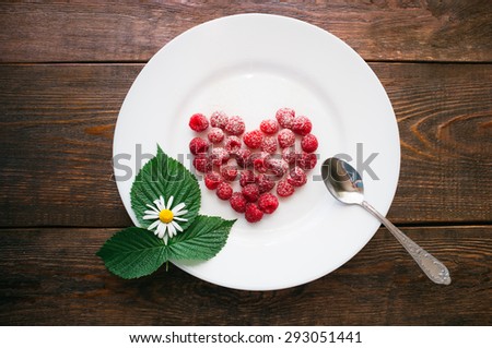 leaves and berries of raspberry lie on a white plate. The red heart from ripe berries powdered with icing sugar. A studio photo on a dark wooden background.