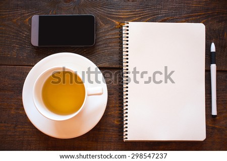 Office business collection: cup of tea, mobile phone, empty notebook and  pen. A photo on a dark wooden table