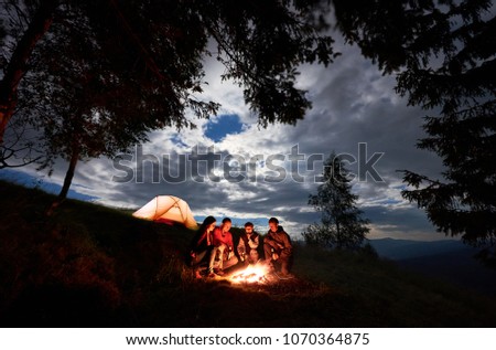 Four person tourists are sitting around a campfire with a beer near the camping and forest at night in the mountains. Blue sky is visible through the clouds. In the distance illuminated orange tent