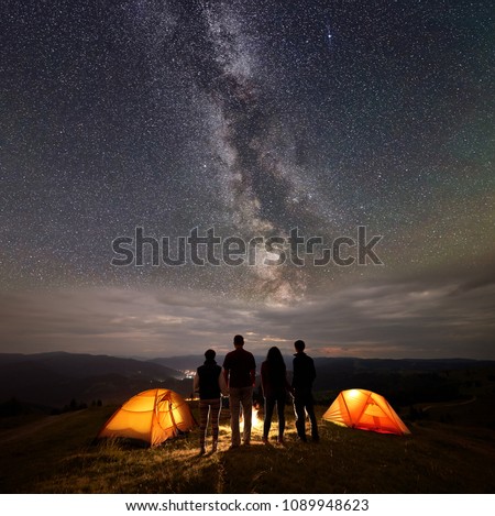 Rear view two young couples hikers holding hands at night near two glowing orange tents, standing by the fire, enjoying starry sky, Milky way, mountains and luminous town. Camping concept at night