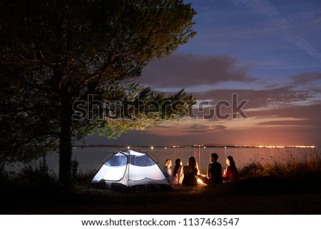 Group of five travellers, girls and boy having a rest on lake shore under tree around campfire at tourist tent on quiet water surface and blue evening sky background. Tourism and camping concept.