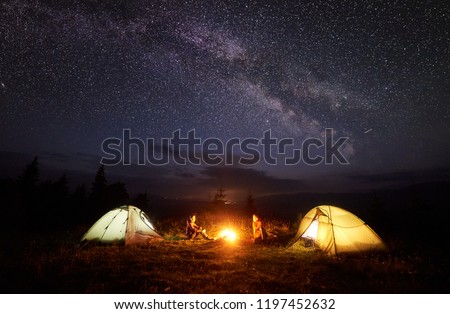 Camping in mountains at night. Bright bonfire burning between two hikers, boy and girl sitting opposite each other near illuminated tents under beautiful evening starry sky and Milky way