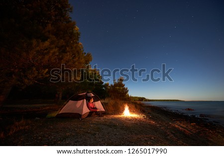 Night camping at sea. Pretty hiker girl resting in entrance of tent near forest at campfire under bright starry sky, enjoying beautiful view of clear blue water. Tourism, active lifestyle concept