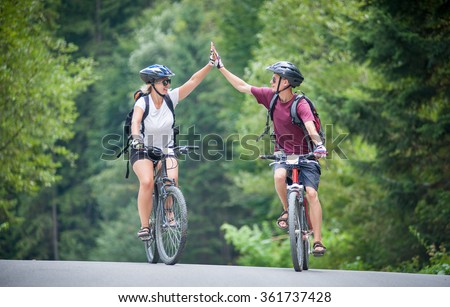 happy couple goes on a mountain asphalt road in the woods on bikes with helmets giving each other a high five