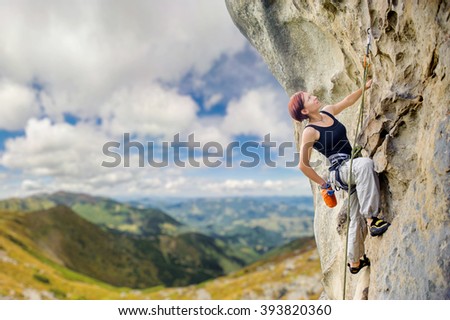 Young athletic female rock climber climbing cliff wall, hanging on one hand and holding hand in magnesium bag. Mountains and blue sky on the background. Copy space on the left.