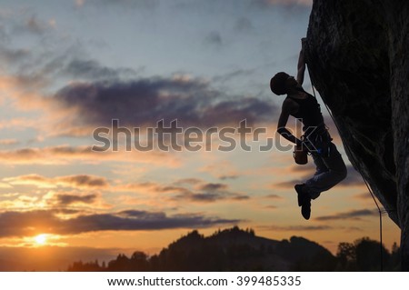 Silhouette of athletic female rock climber climbing steep rock wall against amazing sunset sky in the mountains. Girl is hanging on one hand and holding hand in magnesium bag.