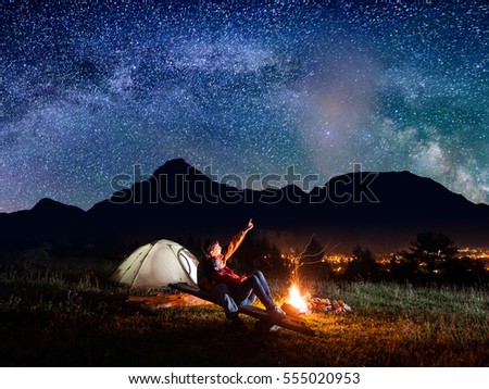 Guy shows his lover in the incredible view of starry sky and Milky way. Pair sitting on boards near campfire on the background high mountains and luminous town