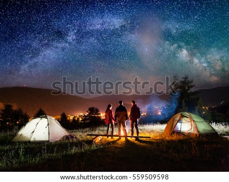 Rear view couple hikers holding hands and their friend stands near campfire and looks at it under night sky with stars on the background mountains and luminous town