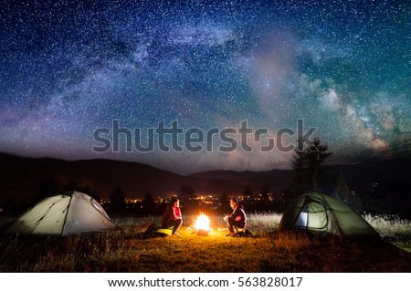 Romantic couple sitting at a campfire near tents in the night under incredibly beautiful starry sky and Milky way. Night camping