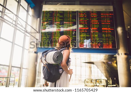Theme travel public transport. young woman standing with back in dress and hat behind backpack and camping equipment for sleeping, insulating mat looks schedule on scoreboard airport station sunny day