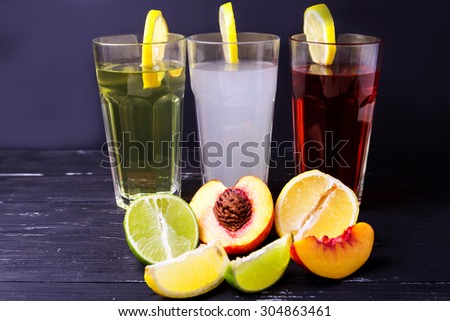 Three fruits soft-drinks with lemon on a edge of a glass. Lime, lemon and peach on a bottom halves and slices on a dark background.