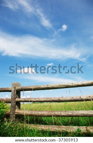 a rail fence in front of a green pasture with a beautiful blue sky with wispy clouds
