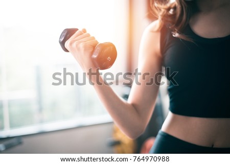 Hand of sports woman lifting dumbbell for weight training near window by right hand for pumping biceps muscle. Workout and Body build up concept. Indoor exercise gym and Sports club theme.