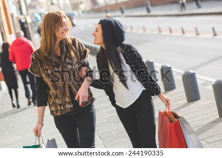 Happy Women Walking in the City with Shopping Bags. One is Holding the Arm of the Other and Looking at Her face Smiling.