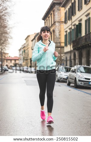 Beautiful Young Woman Jogging Alone on City Street. There is no traffic, early morning and cloudy day. She wears light blue sweatshirt, black leggings and pink shoes. Wearable technology theme.