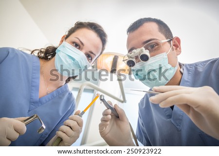 Cheerful Dentists Holding Dental Tools Looking at Camera. Personal or Patient Point of View, POV. They are holding Drill, Mirror, Aspirator and Spreader. Dentist is wearing Dental Magnifying Glasses.