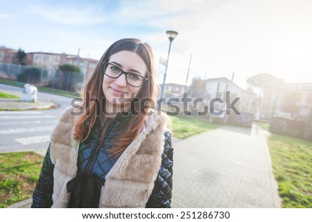Young Woman Portrait at Park in the Morning. The Girl has a Piercing at upper lip and is Looking at Camera. The Park is in a Residential Area, there are some Houses on Background.