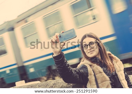Girl Taking Selfie with Train Passing on Background. The Girl is Looking at Smart Phone Camera next to a Rail Crossing; the Train behind is Blurred