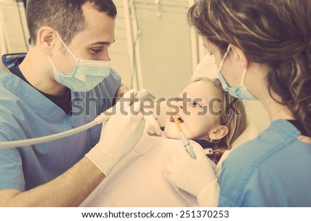 Dentist and Dental Assistant examining Young Girl teeth. Dentist is a Male, Assistant and Patient are Females. Patient is a Young Girl, she\'s Smiling and not scared of Dentist.
