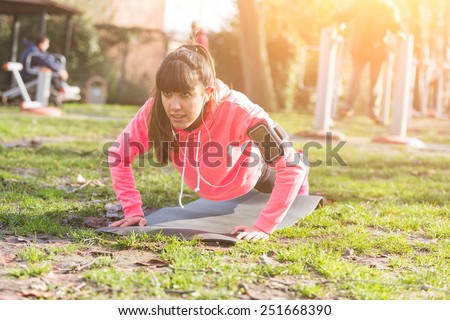 Young Woman Doing Push-Ups Exercises at Park. She is also Listening Music with Earphones. The girl is using a Fitness Mat. Some People on Background doing Exercises too.