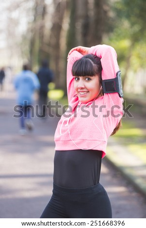 Young Woman Doing Arms and Shoulders Stretching Exercises. She Has a Smart Phone Holder on the Left Arm and she is Listening Music with Earphones. Urban Park setting.