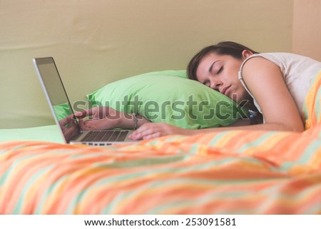 Young Woman Falling Asleep while Using Notebook on Bed. She is Alone. The bed has green and orange Sheets. She still Have her Hands on Keyboard. Technology or Internet addiction theme.