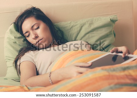 Young Woman Falling Asleep while Using Digital Tablet on Bed. She is Alone. The bed has green and orange Sheets. She still Hold the Tablet with her Hands. Technology or Internet Addiction Theme.