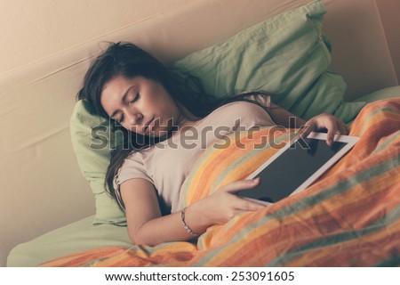 Young Woman Falling Asleep while Using Digital Tablet on Bed. She is Alone. The bed has green and orange Sheets. She still Hold the Tablet with her Hands. Technology or Internet Addiction Theme.