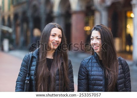 Female Twins Walking on Inner City Street. They wear Black Jackets and Jeans. They Walk side by side Talking and Looking Around. Old Buildings and Narrow Street on Background