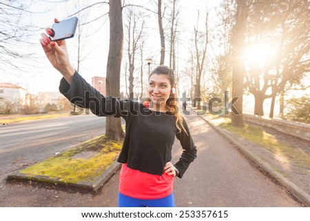 Young Sporty Woman Taking a Selfie at Park. She is Looking at Smart Phone, Wearing a Black Sweatshirt. She has a Smart Phone Holder on her Arm and Listen Music with Earphones.