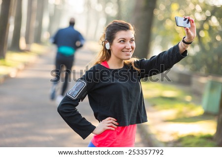 Young Sporty Woman Taking a Selfie at Park. She is Looking at Smart Phone, Wearing a Black Sweatshirt. She has a Smart Phone Holder on her Arm and Listen Music with Earphones.