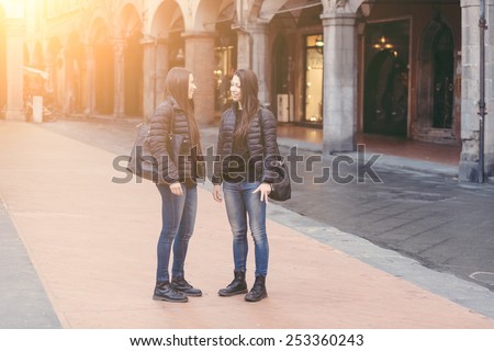 Female Twins Talking Each Other at City Square. They wear Black Jackets. One is Showing her back, the other one is Looking at Her Turned Toward Us.