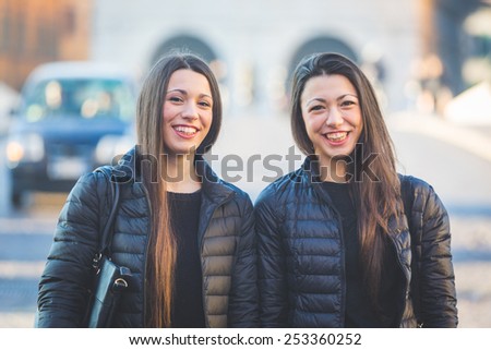 Female Twins Portrait in the City with Traffic on Background. They wear Black Jackets and Long Dark Hair down. There are some Blurred Cars and People on the Street on Background.