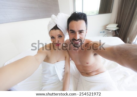 Couple Taking Selfie after Shower. They are in a Modern Hotel Room, They wear white towels and the Woman also has a towel around her Head. They hold the Smart Phone together and Look at it.