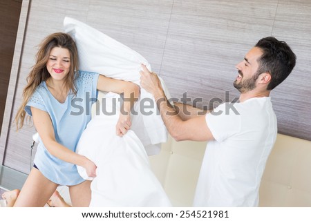Happy Couple Having Pillow Fight in Hotel Room. They Wear Underwear and stand on bendend Knees on the Bed. Everyone is Holding a Pillow. The Woman has Long Blonde Hair, the Man has Black Short Hair.