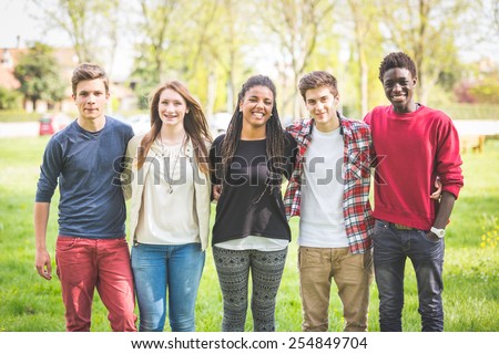 Multiethnic group of teenagers outdoor. They are embraced at park, two boys and one girl are caucasian, one boy and one girl are black. Friendship, immigration, integration and multicultural concepts.
