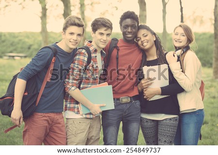 Group of multiethnic teenage students embraced together at park. Two boys and one girl are caucasian, one boy and one girl are black. Friendship, immigration, integration and multicultural concepts.