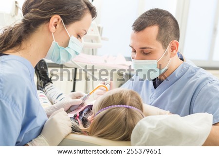 Dentist and dental assistant examining young girl teeth. Dentist is a male, assistant and patient are females. Patient is a young girl, she\'s smiling and not scared of dentist.